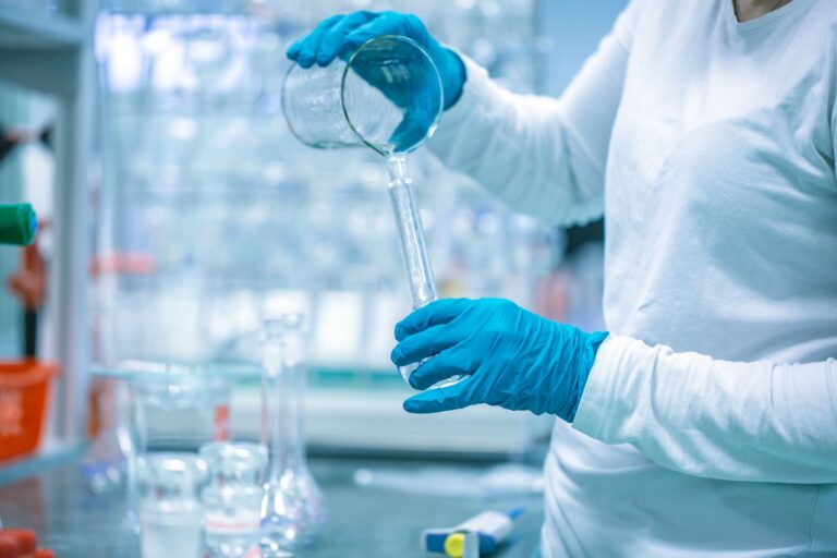 Lab worker wearing all white clothes and blue gloves pouring a clear liquid from a wide container into a thin beaker.
